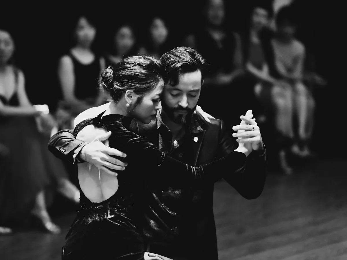 A black-and-white photograph of a tango couple dancing close together in front of a seated audience (representative photograph, not this show's own company)